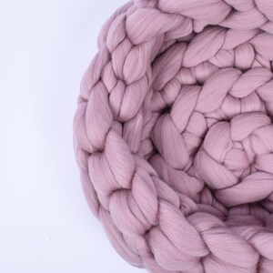May include: A close-up of a large, chunky knit blanket in a soft pink color. The blanket is made of thick yarn and has a braided texture.