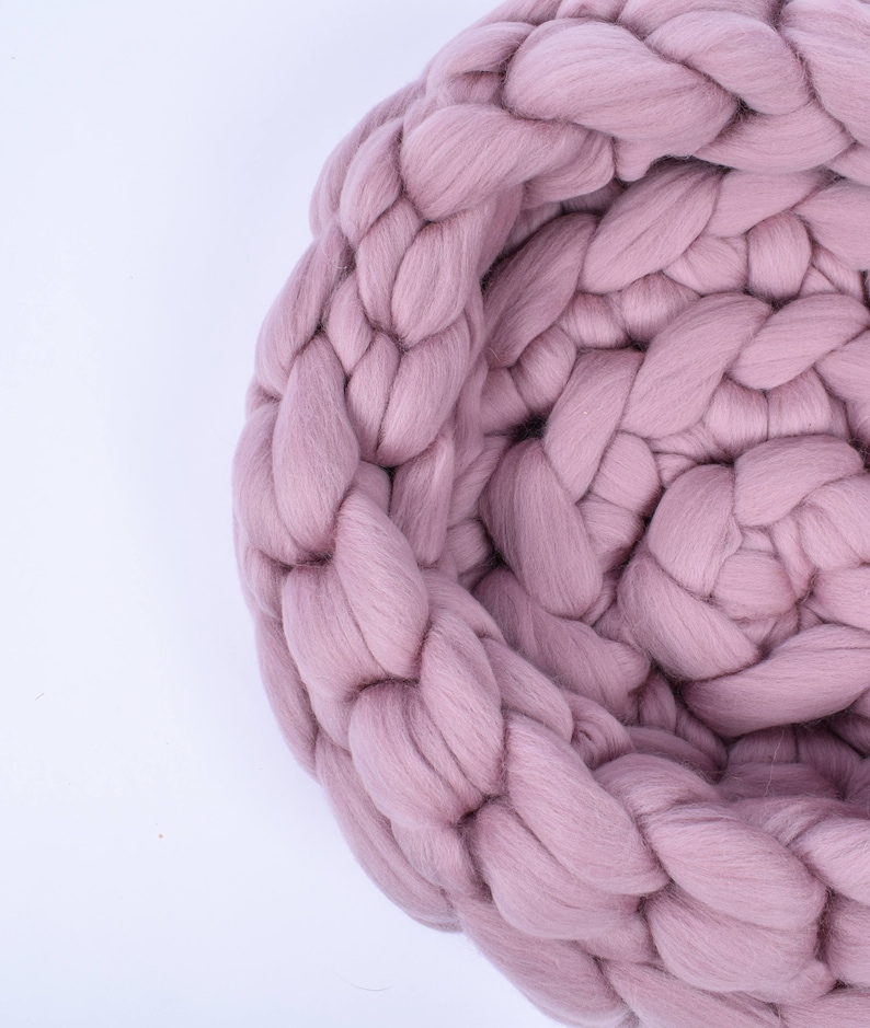May include: A close-up of a large, chunky knit blanket in a soft pink color. The blanket is made of thick yarn and has a braided texture.