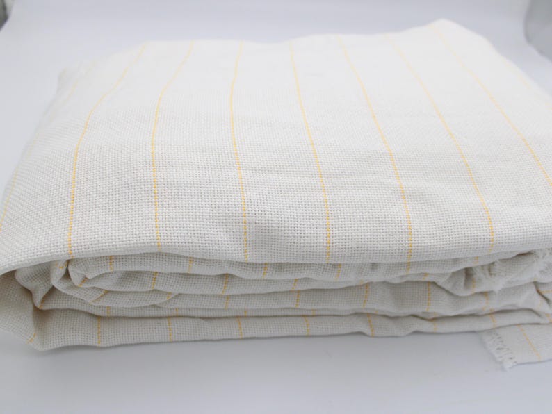 May include: Folded white fabric with vertical yellow stripes. The fabric appears to be a woven material, possibly linen or cotton. The stripes are evenly spaced and add a subtle design element to the textile.