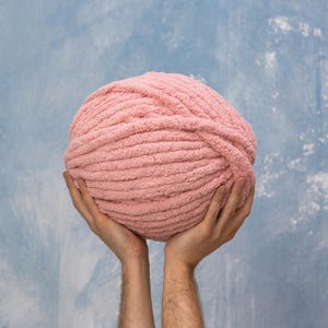 K&ouml;nnte beinhalten: Eine gro&szlig;e Kugel rosa Garn, die in zwei H&auml;nden vor einem blauen Hintergrund gehalten wird.
