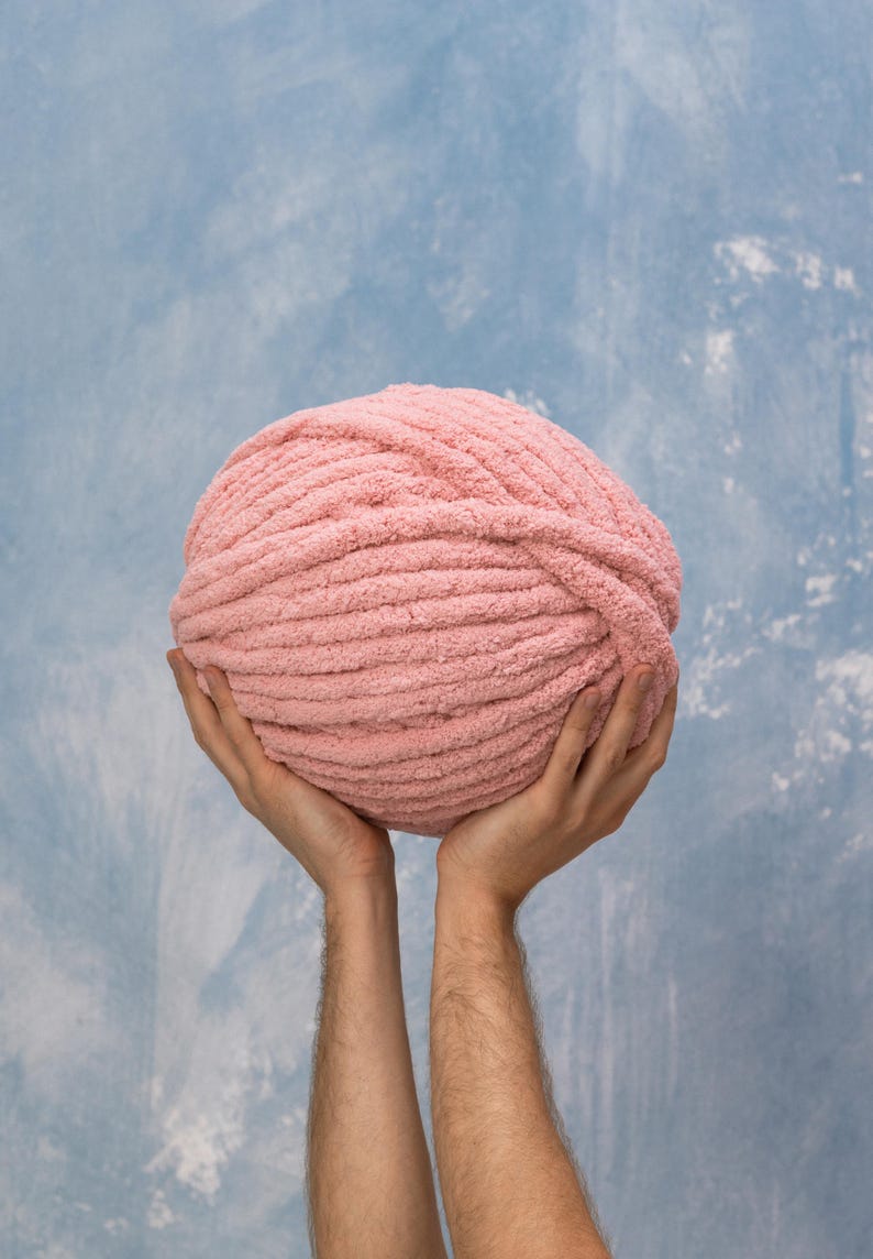 K&ouml;nnte beinhalten: Eine gro&szlig;e Kugel rosa Garn, die in zwei H&auml;nden vor einem blauen Hintergrund gehalten wird.