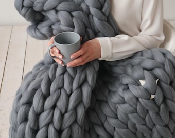 May include: A person is wrapped in a large, grey chunky knit blanket while holding a grey ceramic mug.