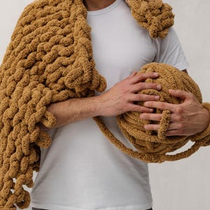 A person holding a large, chunky knit blanket and a ball of matching yarn. The blanket is draped over the shoulder, and the yarn is a warm, golden brown color. The blanket appears soft and cozy.