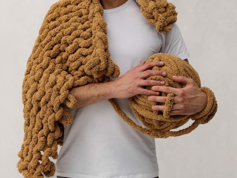 A person holding a large, chunky knit blanket and a ball of matching yarn. The blanket is draped over the shoulder, and the yarn is a warm, golden brown color. The blanket appears soft and cozy.