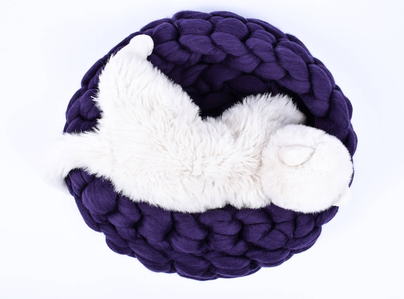 May include: A white fluffy cat curled up in a deep purple knitted pet bed.