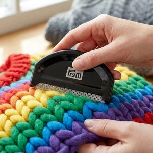 May include: A black Prym fabric shaver with a curved handle and a metallic mesh head, positioned on a thick, chunky knit textile featuring horizontal stripes of red, orange, yellow, green, blue, and purple yarn, used for removing lint and pilling.