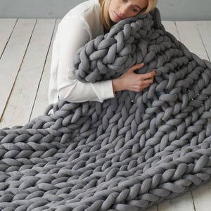 Peut inclure: Une grande couverture en tricot &eacute;pais de couleur gris, faite de laine &eacute;paisse. La couverture est pli&eacute;e et drap&eacute;e sur une personne assise sur un sol en bois.