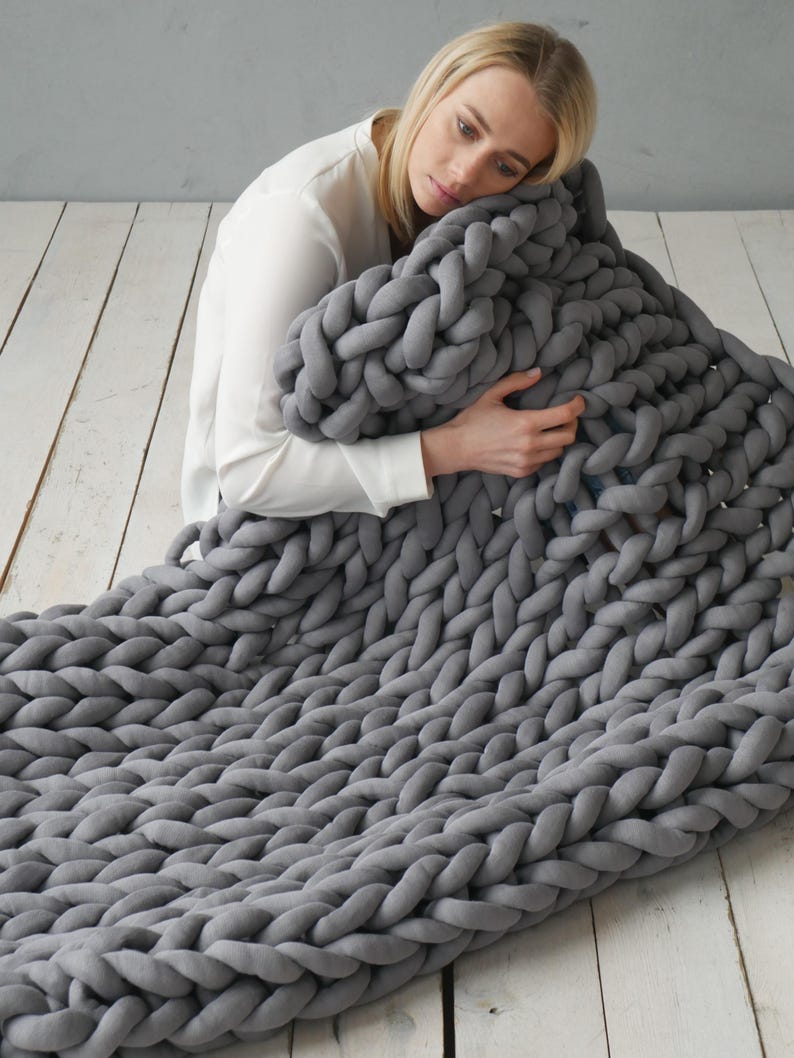 Peut inclure: Une grande couverture en tricot &eacute;pais de couleur gris, faite de laine &eacute;paisse. La couverture est pli&eacute;e et drap&eacute;e sur une personne assise sur un sol en bois.