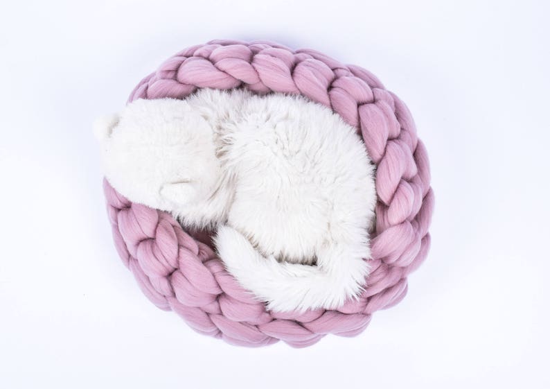 May include: A white cat curled up and sleeping in a pink knitted pet bed.