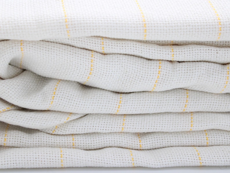 May include: Close-up of a stack of folded, off-white waffle-weave fabric with thin, vertical yellow stripes. The fabric is soft and textured, with visible woven details. The layers create a sense of depth and volume.