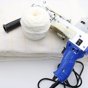 May include: A white and blue electric tufting gun with a black handle and a white yarn ball. The gun is on top of a white fabric with yellow stripes. A black handle and a black cord are also visible.