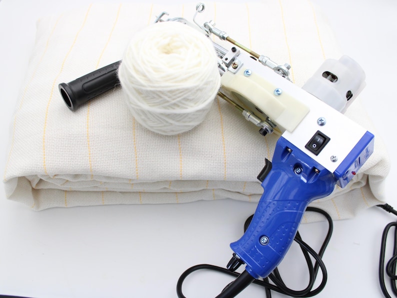 May include: A white and blue electric tufting gun with a black handle and a white yarn ball. The gun is on top of a white fabric with yellow stripes. A black handle and a black cord are also visible.