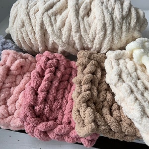 K&ouml;nnte beinhalten: F&uuml;nf verschiedene Farben von dickem Strickgarn, darunter Grau, Rosa, Beige, Braun und Wei&szlig;. Das Garn ist in einer Reihe angeordnet, wobei das wei&szlig;e Garn oben und das graue Garn unten liegt.