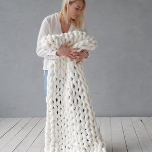 Peut inclure: Une personne tient une couverture en tricot &eacute;pais de couleur blanche. La couverture est faite de fil &eacute;pais et a une texture douce et moelleuse.