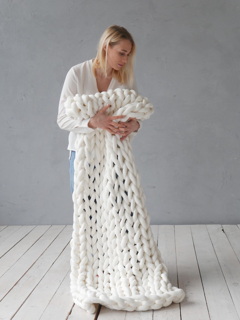 Peut inclure: Une personne tient une couverture en tricot &eacute;pais de couleur blanche. La couverture est faite de fil &eacute;pais et a une texture douce et moelleuse.