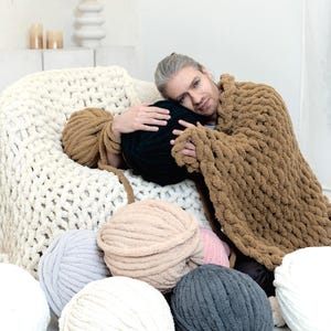 Man is sitting around chunky chenille yarns and holding chunky knit blanket in teddy bear, on the chair cream chenille blanket is laying