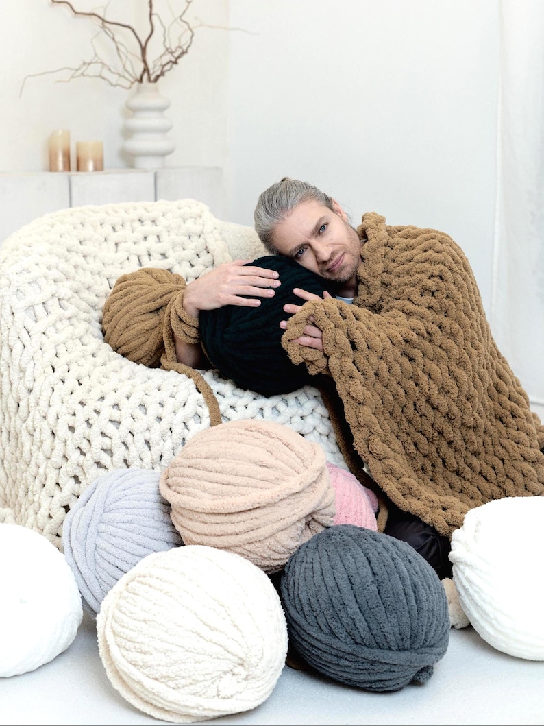 Man is sitting around chunky chenille yarns and holding chunky knit blanket in teddy bear, on the chair cream chenille blanket is laying