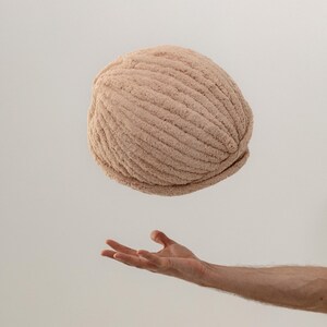 A large ball of beige yarn, suspended in mid-air. The yarn has a soft, textured appearance, with visible ridges. The image shows a hand reaching up towards the yarn, suggesting a crafting or knitting project.
