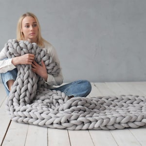 Peut inclure: Une couverture en tricot &eacute;pais de couleur grise avec une personne assise sur un sol en bois blanc tenant la couverture.