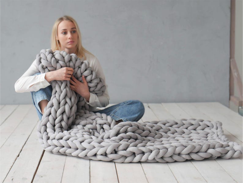 Peut inclure: Une couverture en tricot &eacute;pais de couleur grise avec une personne assise sur un sol en bois blanc tenant la couverture.