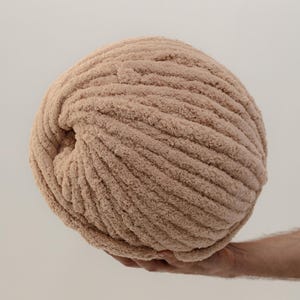 A large ball of beige yarn, held in a hand, is the focal point. The yarn has a thick, textured appearance with visible ridges. The neutral color and soft texture suggest it's suitable for cozy knitting or crochet projects.