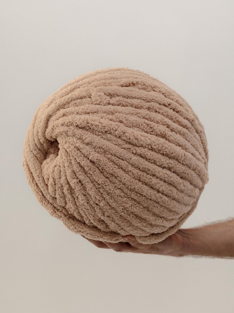 A large ball of beige yarn, held in a hand, is the focal point. The yarn has a thick, textured appearance with visible ridges. The neutral color and soft texture suggest it's suitable for cozy knitting or crochet projects.