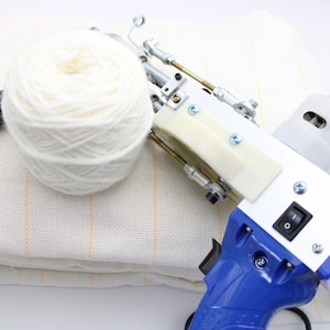 A white and blue tufting gun with a white yarn ball and a piece of fabric. The gun has a blue handle, a white body, and a black switch. The yarn ball is large and fluffy.
