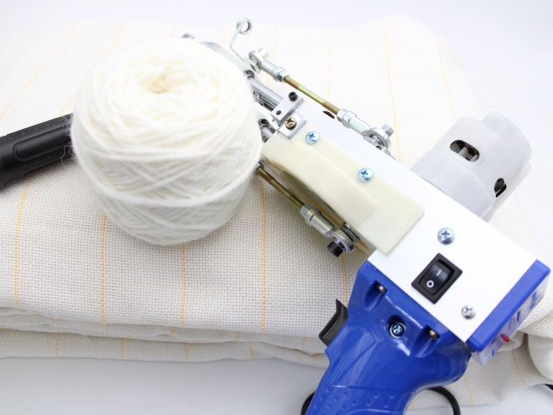 A white and blue tufting gun with a white yarn ball and a piece of fabric. The gun has a blue handle, a white body, and a black switch. The yarn ball is large and fluffy.