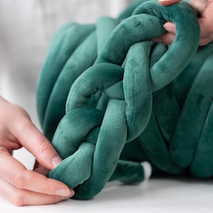 A young woman holding thick, plush velvet yarn, demonstrating the arm knitting technique to create a handmade chunky knit blanket. The cozy, oversized stitches of the giant knit throw are visible as she carefully loops the soft yarn, crafting a warm and stylish home décor piece.