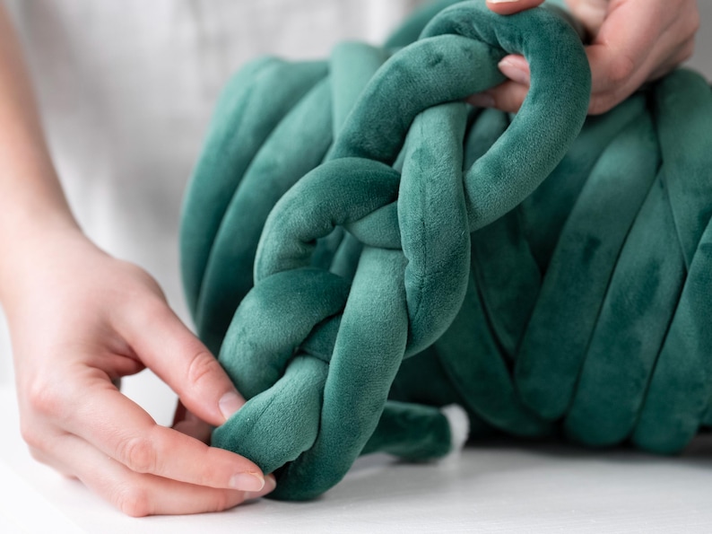 A young woman holding thick, plush velvet yarn, demonstrating the arm knitting technique to create a handmade chunky knit blanket. The cozy, oversized stitches of the giant knit throw are visible as she carefully loops the soft yarn, crafting a warm and stylish home décor piece.