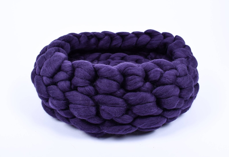 May include: A deep purple knitted pet bed made of thick yarn. The bed is round and has a deep well for a pet to curl up in.