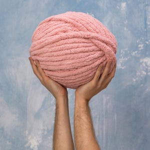 A large ball of soft, light pink yarn held up against a blue and white mottled background. The yarn appears to be thick and textured, ideal for knitting or crocheting projects. The ball is round and full.