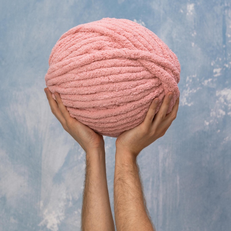 A large ball of soft, light pink yarn held up against a blue and white mottled background. The yarn appears to be thick and textured, ideal for knitting or crocheting projects. The ball is round and full.
