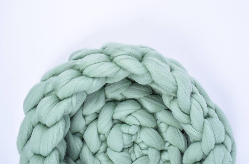 May include: A large, chunky knit blanket in a soft sage green color. The blanket is made of thick, fluffy yarn and is woven in a braided pattern.