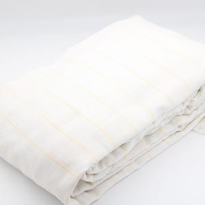 May include: Folded white linen sheet with thin yellow stripes. The sheet is neatly folded, showcasing the fabric's texture and pattern. The neutral color palette and simple design suggest a focus on comfort and understated elegance.
