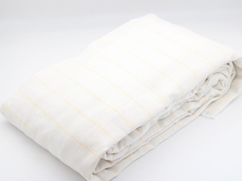 May include: Folded white linen sheet with thin yellow stripes. The sheet is neatly folded, showcasing the fabric's texture and pattern. The neutral color palette and simple design suggest a focus on comfort and understated elegance.