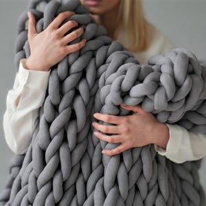 Peut inclure: Une personne tenant une grande couverture en tricot &eacute;pais de couleur grise. La couverture est faite de fil &eacute;pais et doux et a une trame l&acirc;che et ouverte.