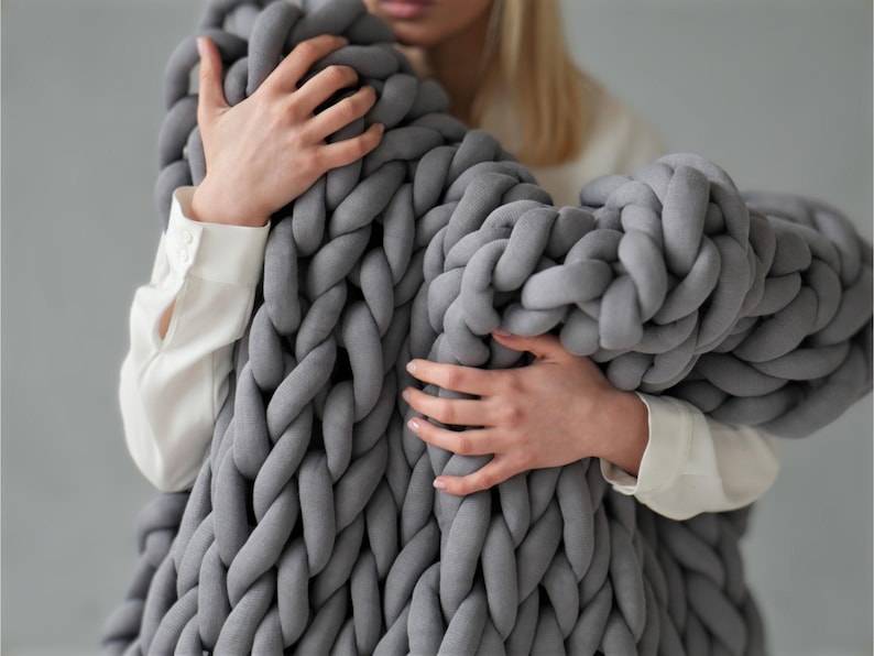 Peut inclure: Une personne tenant une grande couverture en tricot &eacute;pais de couleur grise. La couverture est faite de fil &eacute;pais et doux et a une trame l&acirc;che et ouverte.
