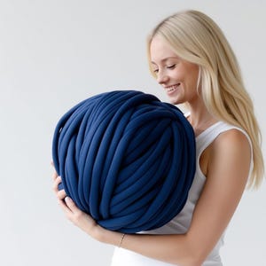 Puede incluir: Una gran bola redonda de hilo azul marino. El hilo es grueso y parece estar hecho de un material suave y tejido. La bola la sostiene una persona que lleva una camiseta sin mangas blanca.