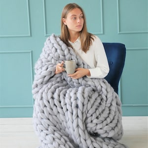 May include: A woman sits in a blue chair, wrapped in a large, chunky knit gray blanket. She holds a gray mug. The blanket is made of thick, soft yarn, with large, visible stitches. The background is a light blue wall.