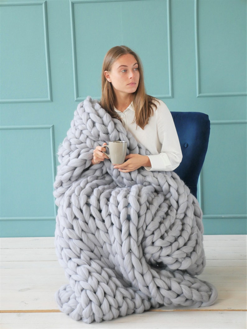 May include: A woman sits in a blue chair, wrapped in a large, chunky knit gray blanket. She holds a gray mug. The blanket is made of thick, soft yarn, with large, visible stitches. The background is a light blue wall.