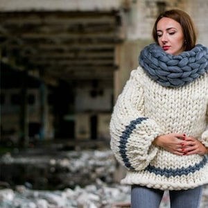 May include: A white chunky knit sweater with gray stripes on the sleeves and a gray chunky knit scarf. The sweater has a loose fit and a turtleneck.