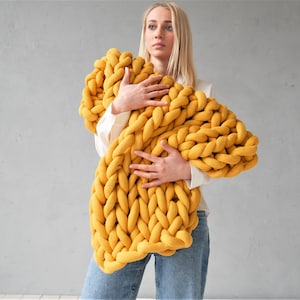 以下が含まれることがあります： マスタードイエローの大きな厚手のニットブランケットを持っている人。ブランケットは厚手の柔らかな糸でできており、リラックスして居心地の良い雰囲気です。