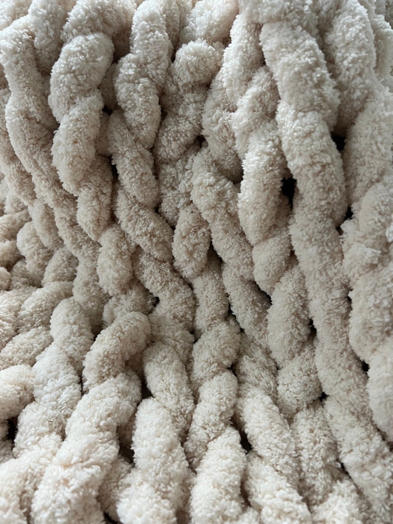 K&ouml;nnte beinhalten: Nahaufnahme einer cremefarbenen, grob gestrickten Decke. Die Decke besteht aus dickem, flauschigem Garn und hat eine weiche, strukturierte Oberfl&auml;che.