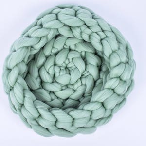 May include: A large, sage green, chunky knit throw blanket is woven in a braided pattern. The blanket is rolled up into a circle, creating a nest-like shape.