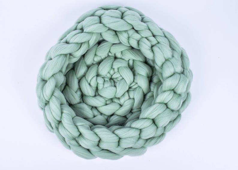 May include: A large, sage green, chunky knit throw blanket is woven in a braided pattern. The blanket is rolled up into a circle, creating a nest-like shape.