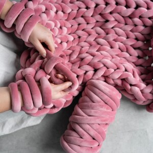 A young woman holding thick, plush velvet yarn, demonstrating the arm knitting technique to create a handmade chunky knit blanket. The cozy, oversized stitches of the giant knit throw are visible as she carefully loops the soft yarn, crafting a warm and stylish home décor piece.