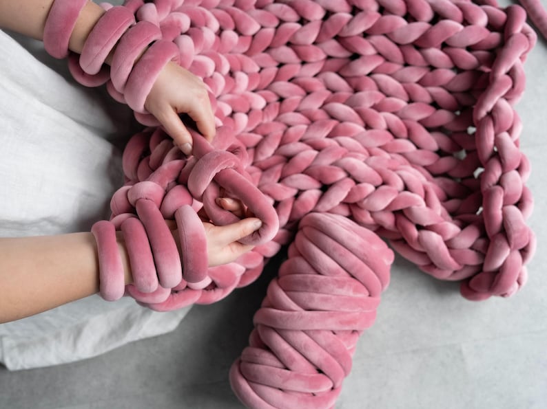 A young woman holding thick, plush velvet yarn, demonstrating the arm knitting technique to create a handmade chunky knit blanket. The cozy, oversized stitches of the giant knit throw are visible as she carefully loops the soft yarn, crafting a warm and stylish home décor piece.