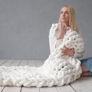Peut inclure: Une couverture en tricot &eacute;pais de couleur blanche avec une personne assise dessus. La couverture est faite de fil &eacute;pais et a une texture douce et confortable.