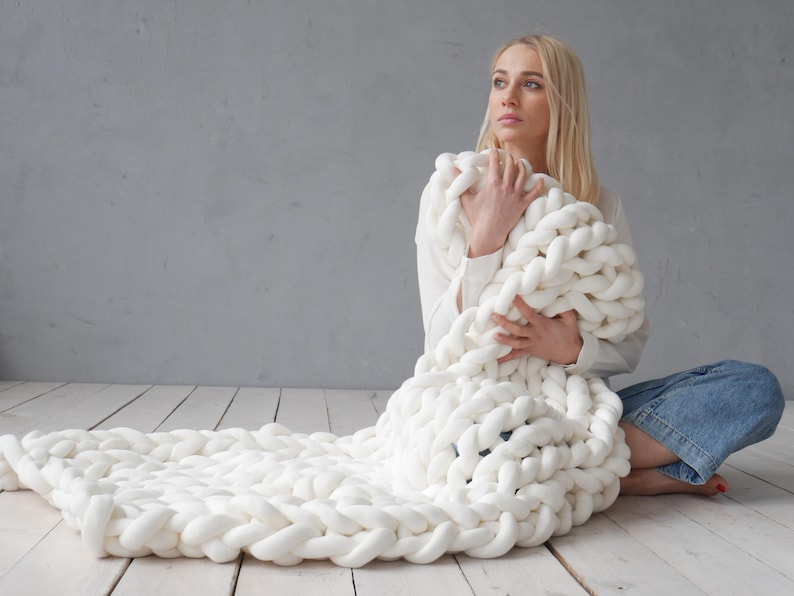 Peut inclure: Une couverture en tricot &eacute;pais de couleur blanche avec une personne assise dessus. La couverture est faite de fil &eacute;pais et a une texture douce et confortable.